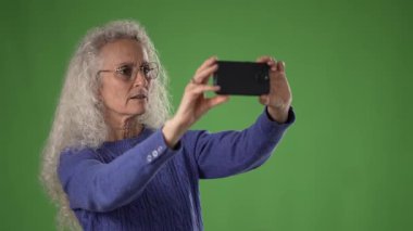50 'li yaşlarda, uzun gri saçlı olgun bir kadın canlı yeşil bir ekranın önünde beklerken akıllı telefonunu tutar ve selfie çeker..