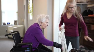 Yaşlı bir kadın tekerlekli sandalyede otururken, başka bir kadın ona kıyafet seçmesinde yardım ediyor. Parlak bir odadalar ve yakınlarda günlük giysiler var..