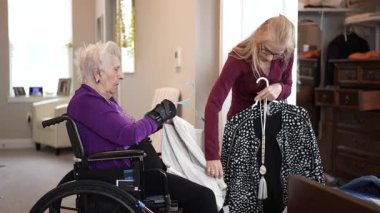 Bir bakıcı, tekerlekli sandalyede oturan yaşlı bir kadına kıyafet seçmesinde yardımcı olur. Ortam rahat ve iyi aydınlatılmış, destekleyici atmosferi geliştiriyor..