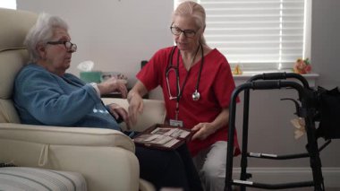 80 ya da 90 yaşlarında yaşlı bir kadın koltukta otururken, kırmızı üniformalı bir sağlık görevlisi konforlu bir evde bakım ve destek sağlıyor..