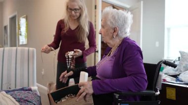 Samimi bir iç mekanda, tekerlekli sandalyedeki olgun bir kadın çeşitli mücevher parçalarını paylaşan genç bir kadınla heyecan verici bir şekilde yakınlaşıyor..