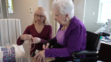 Sadık bir bakıcı, tekerlekli sandalyedeki olgun bir kadına yardım eder ve kolye takar. Sıcak, rahatlatıcı atmosfer güçlü bağlarını ve ilgilerini vurguluyor..