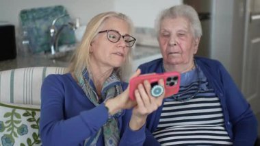 Bir kız, evde rahat bir ortamda yaşlı annesine fotoğraf göstermek için akıllı telefonunu kullanır. Birlikte anıları paylaşıyorlar, sevgiyi ve özeni vurguluyorlar..