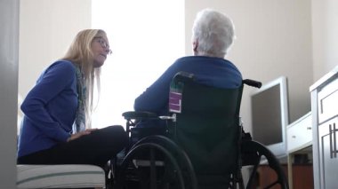 Doksanlı yaşlarında bir kadın tekerlekli sandalyede oturuyor ve kızından samimi bir ilgi görüyor. Kapalı alan ayarları yakın bağlarını vurguluyor..