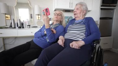 Bir kız, evde rahat bir şekilde otururken akıllı telefon kullanarak, birbirleriyle olan bağlarını ve ilgilerini göstererek, yaşlı annesiyle neşeli anlar paylaşır..