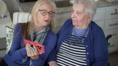 Kapalı bir ortamda, 60 'lı yaşlardaki bir kadın yaşlı annesinin fotoğraflarını akıllı bir telefondan gösteriyor, ziyaretleri sırasında sevgi ve bağlantı anlarını yakalıyor..