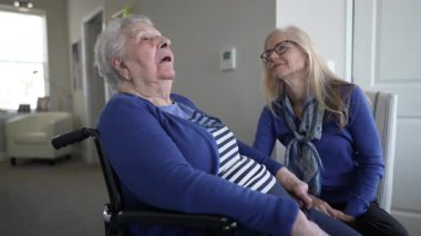 Kızı evde duygusal destek sağlarken 90 'larında yaşlı bir kadın tekerlekli sandalyede oturuyor. Sıcak bir ortamda bir anlık birlikteliği paylaşıyorlar..