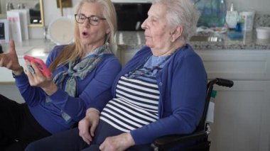 90 'lı yaşlarda bir kadın tekerlekli sandalyede otururken, kızı ona akıllı telefondan bir şey gösteriyor, ev ortamında neşeli anılarını paylaşıyor..