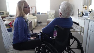 Sıcak, kapalı bir mekanda, bir kadın yaşlı annesine yakın bir tekerlekli sandalyede oturuyor, aile ziyareti sırasında bir anlık ilgi ve bağlantı paylaşıyor..