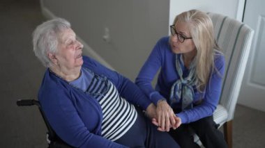 Tekerlekli sandalyedeki yaşlı bir kadın ziyaret sırasında kızından sevgi dolu destek alır. Sıcak, kapalı bir mekanda birlikte hassas bir anı paylaşıyorlar..