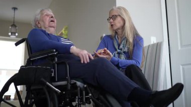 Tekerlekli sandalyedeki yaşlı bir kadın kızıyla sıcak bir ziyaretten hoşlanıyor. Sıcak ev ortamlarında sevgi ve bakım anlarını paylaşıyorlar..