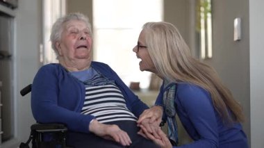 Bir kız, evde yaşlı annesine tekerlekli sandalyede yardım ediyor. Ev ortamındaki günlük aktivitelerinde sevgi ve ilgi gösteriyor..