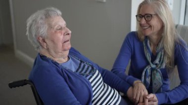 90 'lı yaşlarda yaşlı bir kadın tekerlekli sandalyede otururken mutlu bir şekilde gülümsüyor, sıcak bir anı kızıyla paylaşıyor..