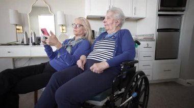 Tekerlekli sandalyedeki yaşlı bir kadın evde kızıyla samimi anlar yaşıyor, akıllı telefon kullanarak bağlı kalıyor ve birlikte anılara değer veriyor..