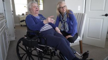 90 'lı yaşlarda yaşlı bir kadın tekerlekli sandalyede oturuyor ve kızıyla evde sohbet ediyor. Atmosfer, ziyaretleri sırasında sıcaklığı ve ilgiyi yansıtıyor..