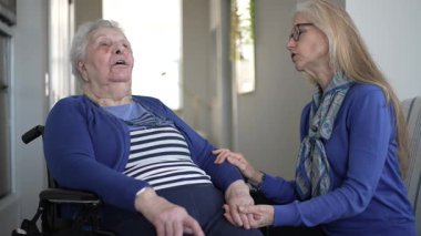 90 'lı yaşlarda yaşlı bir kadın, günlük ziyareti sırasında kızından sevgi dolu bir destek alarak evinde tekerlekli sandalyede oturuyor. İçerideki atmosfer sıcak ve davetkar..