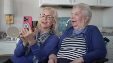 90 'lı yaşlarda bir kadın tekerlekli sandalyede otururken, kızı ona akıllı telefondan bir şey gösteriyor, evde sevgi ve destek anı yakalıyor..