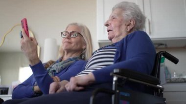 Kapalı bir ortamda, tekerlekli sandalyedeki yaşlı bir kadın akıllı telefon aracılığıyla kızıyla iletişim kuruyor, gülümsemelerini ve güzel anılarını paylaşıyor..