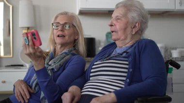Tekerlekli sandalyedeki yaşlı bir kadın, kızı ona akıllı bir telefon gösterirken gülümsüyor. Sıcak bir evde sevgi ve beraberlik anları yakalıyor..