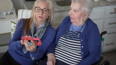 60 'lı yaşlarda bir kadın, 90' lı yaşlardaki yaşlı annesine sıcak bir mutfakta oturup, anılarını paylaşırken ve birlikte vakit geçirirken akıllı telefon kullanmasında yardımcı olur..