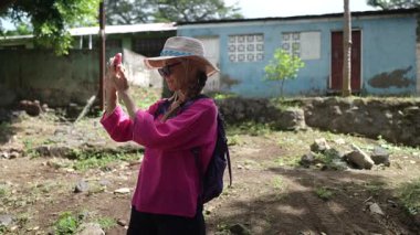 Şapkalı mutlu bir kadın Nikaragua 'daki Ometepe Adası' nın bereketli manzaralarını keşfederken telefonuyla fotoğraf çekiyor. Çevrelerde yerel mimari ve doğa yer alıyor.