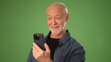 Kıdemli girişimci, arkadaşları ve iş arkadaşlarıyla birlikte canlı yeşil ekran arka planına sahip bir stüdyoda canlı bir atmosfer yaratarak akıllı telefonunda neşeli bir şekilde görüntülü sohbet ediyor..