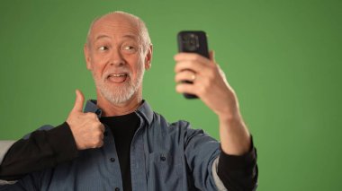 Gri sakallı olgun bir adam gülümsüyor ve yeşil perde arkasında selfie çekiyor. Onun ifadesi mutluluk ve pozitiflik taşır..