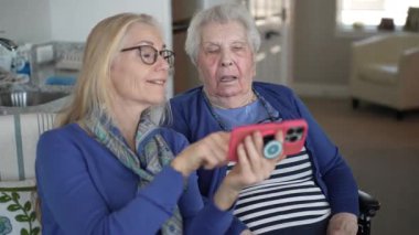 Tekerlekli sandalyedeki yaşlı bir kadın, kızıyla akıllı telefon kullanırken gülümsüyor..