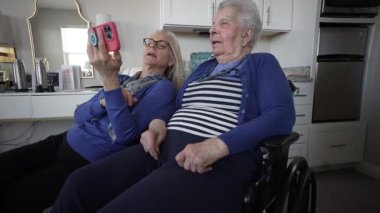 Tekerlekli sandalyede oturan yaşlı bir kadın, evinde sıcak bir ziyaretin tadını çıkarırken akıllı bir telefonla kızıyla birlikte anılarını ve neşesini paylaşıyor..