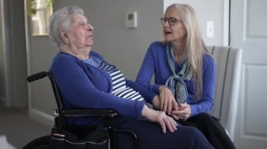 Bir kız çocuğu, eve yaptığı özel bir ziyaret sırasında yaşlı annesine yakın bir tekerlekli sandalyede sevgi ve destek sunuyor. Hava sıcak ve şefkatli..
