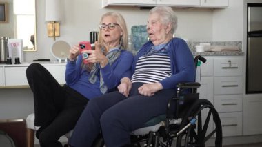 Tekerlekli sandalyedeki yaşlı bir kadın kızıyla mutlu bir şekilde rahat evlerinde akıllı bir telefon kullanarak anılarını ve birlikteliklerini kutluyor..