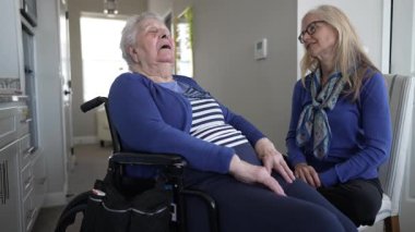 90 'lı yaşlarda bir kadın tekerlekli sandalyede oturuyor, kızı sıcak evlerini ziyaret ederken ona eşlik ederken gülümsüyor..