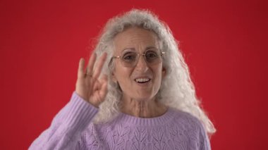 Beyaz saçlı ve gözlüklü orta yaşlı bir kadın dostça bir jest yaparak elini kaldırırken sıcak bir şekilde gülümsüyor. Kıpkırmızı arka plan çekiciliğini ve neşesini arttırıyor..