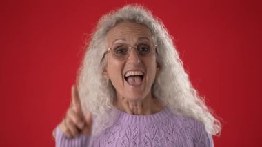 Beyaz saçlı ve gözlüklü orta yaşlı bir kadın çağrı yaparken gülümsüyor. O rahat bir mor süveter giyer ve parlak kırmızı bir arka plan üzerine kuruludur..