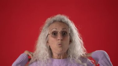 Beyaz saçlı ve gözlüklü orta yaşlı bir kadın heyecandan ellerini kaldırarak gülümsüyor. Yumuşak mor bir kazak giymiş, enerji saçıyor..