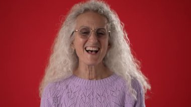Gözlüklü ve kıvırcık beyaz saçlı neşeli orta yaşlı bir kadın, çarpıcı kırmızı bir arka fonun önünde açık mor bir süveter giyerek kahkaha atıyor..