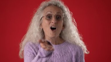 50-60 yaşlarında olgun bir kadın öpüşürken gözlük takar ve mor bir kazak giyer. Parlak kırmızı arkaplan canlı bir karşıtlık ekler.