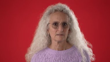 50 'li ya da 60' lı yaşlarda, uzun beyaz saçlı ve gözlüklü olgun bir kadın başını sallıyor. Çarpıcı bir kırmızı arka plana karşı hayır diyor. Yüzünde ciddi bir ifade var..