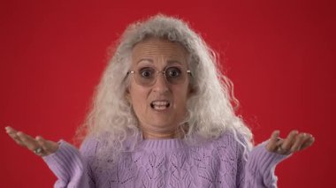 Çekici özellikleri ve gözlüğü olan orta yaşlı bir kadın elleriyle bilmediğim jestler yapıyor. Cesur, kırmızı bir arka plana karşı bir merak veya belirsizlik duygusu taşıyor..