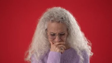 50 ya da 60 yaşlarında, beyaz saçlı ve gözlüklü olgun bir kadın, kırmızı bir fonda açık mor bir süveter giyerken parlak bir fikri parmağıyla tutkuyla işaret ediyor..