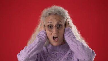 50 ya da 60 yaşlarında olgun bir kadın ifade edici bir görünüm sergilerken kulaklarını kapatır. Kıvırcık beyaz saçları ve gözlüğü var, canlı kırmızı bir arkaplanda..