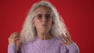 Beyaz saçlı ve gözlüklü orta yaşlı bir kadın mor bir kazak giyerken mutlu bir şekilde onunla olmayı diliyor. Canlı kırmızı arka plan onun neşeli tavrını artırıyor..