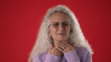 Beyaz saçlı, gözlüklü, boğazı ağrıyan orta yaşlı bir kadın. Mor bir kazak giymiş, parlak kırmızı bir arkaplanda poz veriyor..