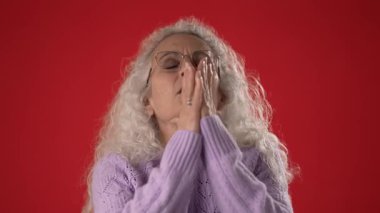 Beyaz saçlı ve gözlüklü orta yaşlı bir kadın ellerini birleştirip hareketli kırmızı bir arka plana karşı duruyor. Zarafet ve cazibe saçıyor..