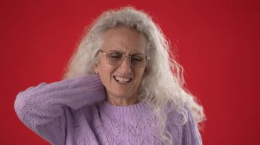 Kıvırcık beyaz saçlı ve gözlüklü orta yaşlı bir kadın koyu kırmızı bir arka plan karşısında rahat bir mor süveter giyerken boyun rahatsızlığı yaşıyor gibi görünüyor..