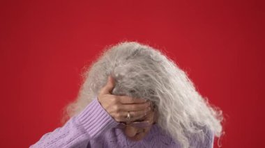 Bukleli beyaz saçlı orta yaşlı bir kadın gözlük ve mor bir kazak giyiyor. Saçlarını parlak kırmızı bir arka planda tutarken başı ağrıyor..