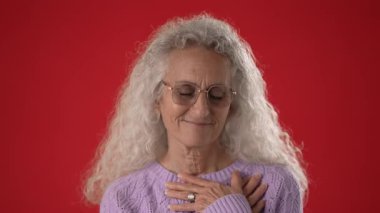 Lavanta kazaklı orta yaşlı bir kadın gülümseyerek göğsüne hafifçe dokunuyor. Uzun beyaz saçları var ve parlak kırmızı bir arka planda gözlük takıyor..