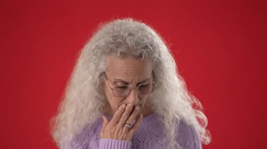 Gözlük takan orta yaşlı bir kadın canlı kırmızı bir arka planın önünde kendinden emin bir şekilde duruyor. Beyaz kıvırcık saçları var ve kameraya şevkle işaret ediyor..