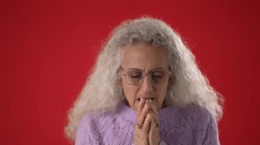 Beyaz saçlı ve gözlüklü orta yaşlı bir kadın ellerini birbirine sıkıca bastırırken hafifçe gülümsüyor. Canlı kırmızı bir zemin ile çevrili..