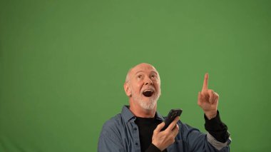 Neşeli bir ifadeye sahip olgun bir adam akıllı telefonunu yukarı doğru tutar, sanki yeşil bir arkaplan önünde heyecan verici haberleri paylaşır gibi..